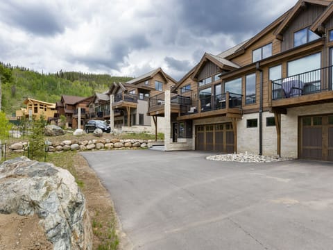 A row of modern, rustic-style homes with large windows and multiple garages, set on a sloped, landscaped area with a large driveway and cloudy skies overhead.