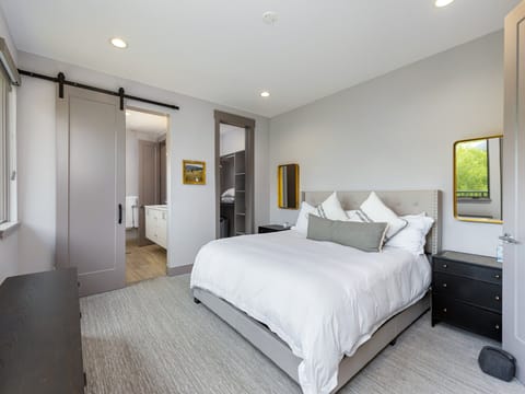 A bedroom with a large bed with white bedding, a nightstand on each side, a sliding door leading to an en-suite bathroom, a dresser, and a window with natural light.