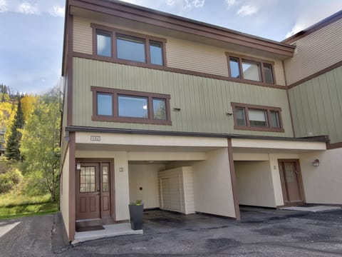 A two-story townhouse with brown trim, an attached covered carport, and green and beige exterior, located in a wooded area. The front door is on the left under the covered area, marked 2570.