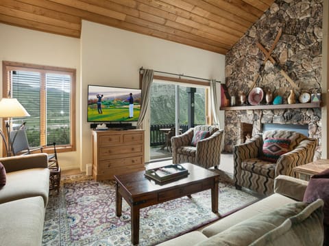 A cozy living room with a stone fireplace, wooden ceiling, and large window overlooking mountains. Two armchairs face a TV showing a golf scene. A coffee table and sofa complete the setting.