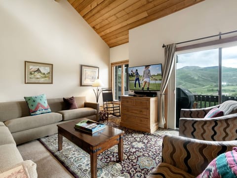 A living room with a high, wooden ceiling, a beige sectional sofa, armchairs, a wooden coffee table, a TV, landscape paintings, and a glass door opening to a balcony with a view of green hills.