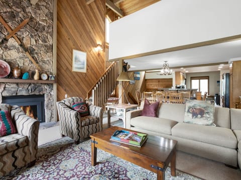 A cozy living room with a beige sofa, patterned chairs, a wooden coffee table, and a fireplace. Wooden stairs lead to the upper floor, and the dining area and kitchen are visible in the background.