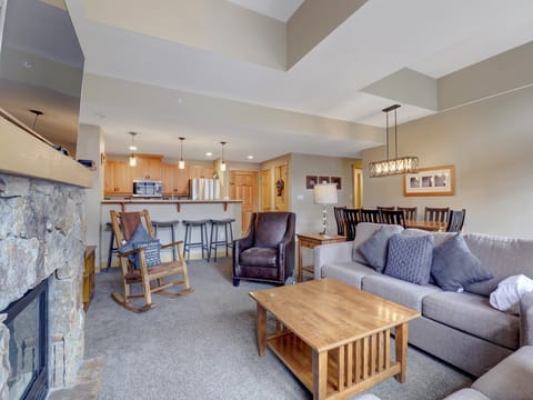 Bright and welcoming great room featuring a plush sofa set, leather armchair, and rustic rocking chair. The open floor plan flows seamlessly to a dining table for six and a full kitchen with a breakfast bar, perfect for gathering after a day on the slopes.
