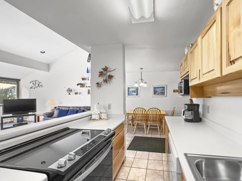 A kitchen interior with a stove and counter in the foreground. Wooden cabinets, a coffee maker, and a dining area with a wooden table and chairs are visible. The living area is in the background.