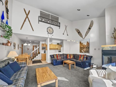 A spacious living room with two sofas, a wooden coffee table, wall-mounted skis and snowshoes, a kitchen in the background, and a staircase leading to an upper level.