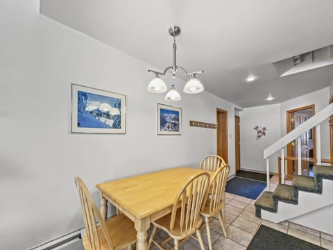 A dining area with a wooden table and four chairs, illuminated by a ceiling light fixture. Two landscape paintings hang on a white wall. A staircase with green carpet and tile flooring is visible.