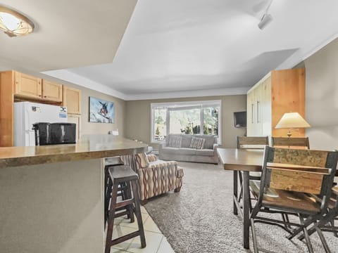 A cozy living area with a kitchen. A countertop with stools, a sofa, armchairs, a dining table with chairs, a window with a scenic view, and a wall-mounted TV are present. Carpeting covers the living area floor.