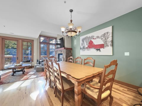 A dining room with a wooden table and six chairs, green walls, a fireplace, a chandelier, a painting of a barn, and large windows opening to a balcony with outdoor scenery.