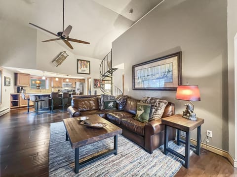A cozy living room features a brown leather sofa with wildlife-themed cushions, wooden tables, and a textured rug. A spiral staircase and open-plan kitchen are visible in the background.