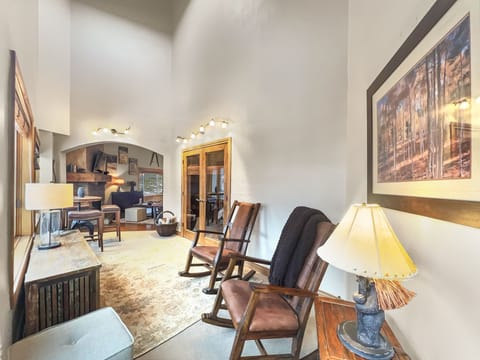 A cozy room features two rocking chairs, a wooden bench, a table lamp, and a framed painting. The room extends to a brighter area with a desk and various furnishings.