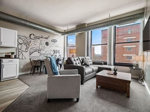 -Bright living room with large industrial windows bringing in natural light   - Cozy sofa and armchair perfect for streaming or relaxing after exploring Cleveland   - Open concept layout with dining table and Cleveland wall art accent