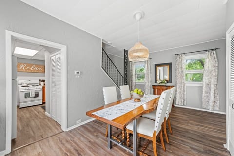 Bright dining area showcases a live-edge wooden table with a lace runner, surrounded by white upholstered chairs and topped with a bowl of green apples. A woven pendant light hangs above, while a row of white louvered closet doors lines the gray wall. Sun