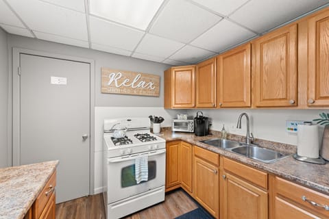 Well-appointed L-shaped kitchen with warm oak cabinets, granite-look countertops, a gas stove, and a double sink under ample cabinet storage. Cozy sign invites you to unwind as you cook and connect. 🍳🏡✨