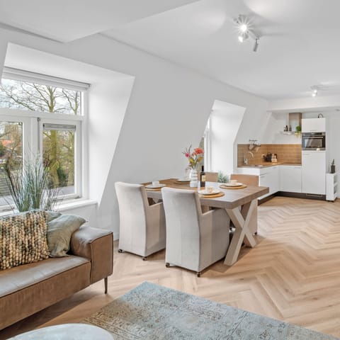 A stylish open-concept interior with a beige sectional, rustic wooden dining table, and large windows showcasing an inviting, airy atmosphere.