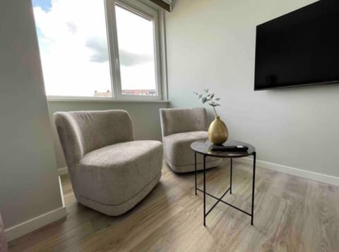 Cozy corner with two beige chairs, a sleek black side table holding a gold vase, large windows for natural light, and a wall-mounted TV for a modern touch.