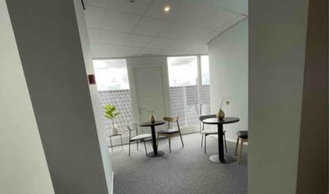 Bright lounge area with round black tables, simple wooden and metal chairs, a potted plant, and large windows offering natural light and rooftop views.