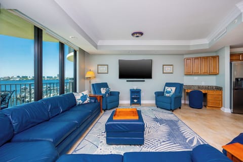 Bright living room featuring a blue sectional sofa, armchairs, TV, and a panoramic view of the bay through the large windows