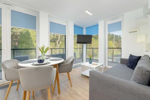 A bright living room with large windows, a round dining table, and modern decor. The room has light wood flooring and a cozy atmosphere.