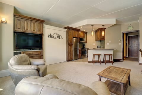 A cozy living area with beige leather chairs, a wooden coffee table, and a wall-mounted TV. A kitchen with wooden cabinets, stainless steel appliances, a breakfast bar with stools, and pendant lights is in the background.