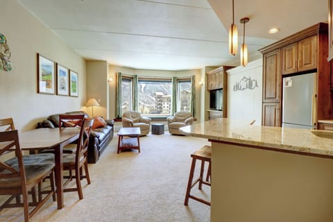 A well-lit open living area with a kitchen counter, dining table, leather couch, armchairs, and large windows showcasing mountain views. The space is modern with wood cabinetry and light-colored walls.