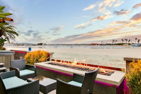 A cozy beachfront patio featuring a modern fire pit, comfortable wicker seating with blue cushions, and seamless access to the indoor living space. Surrounded by lush tropical plants, this inviting outdoor area is perfect for relaxing and enjoying warm ev