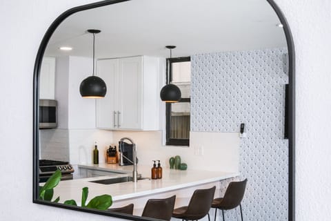 A stylish reflection in a modern arched mirror showcasing a contemporary kitchen with a large island, sleek white cabinetry, and elegant pendant lights. The scene is complemented by bar stools and a subtle patterned accent wall, creating a sophisticated a