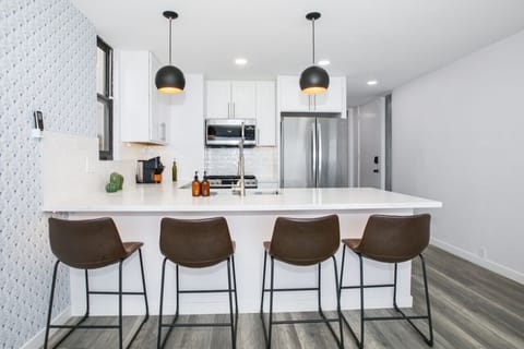 A modern and stylish kitchen featuring a large island with sleek white countertops, complemented by contemporary bar stools and pendant lighting. The kitchen is fully equipped with stainless steel appliances and crisp white cabinetry, creating a functiona