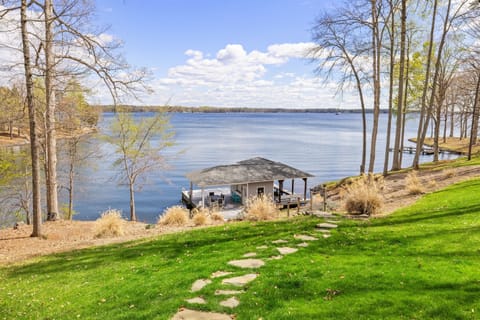 A serene lakeside view with a boat dock and a walking path leading through a grassy lawn flanked by a canvas of trees.