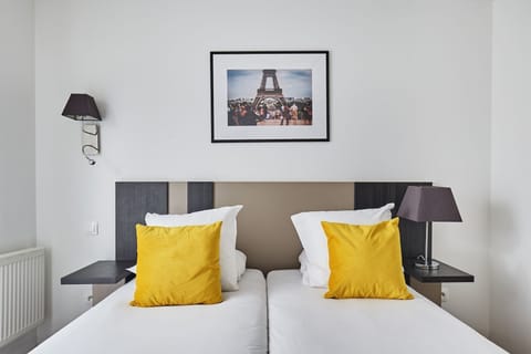 This cozy studio features zip beds adorned with bright yellow pillows, complemented by modern wall sconces and a framed photograph of the Eiffel Tower.