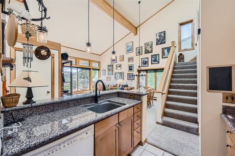 A kitchen with granite countertops, wooden cabinets, and a sink. Stairs lead upstairs on the right, and a wall decorated with framed pictures is visible in the background.