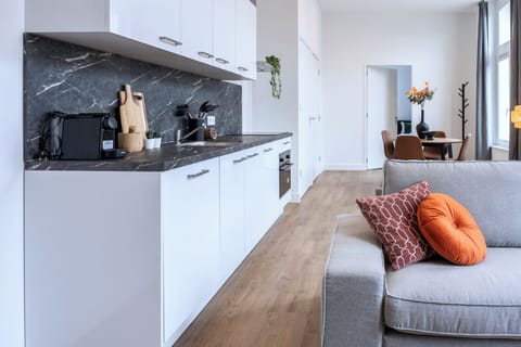 Modern living space with a sleek white kitchen, gray marble backsplash, cozy gray sofa accented with orange pillows, and a dining area featuring vibrant decor.