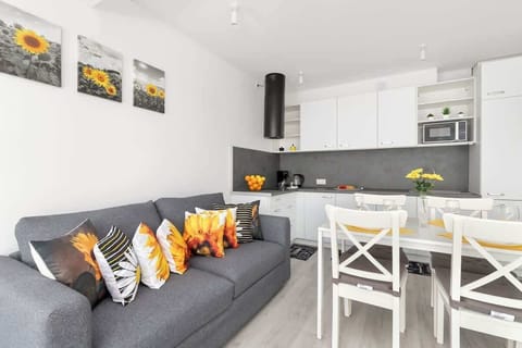 A modern white kitchen with a small dining table, set for a meal with vibrant orange cushions on the chairs. The room is bright and well-organized with overhead cabinets.