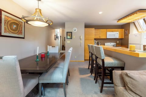 A modern open-plan kitchen and dining area. The dining table is set for three with candles, and the kitchen features a wooden counter with four upholstered barstools.