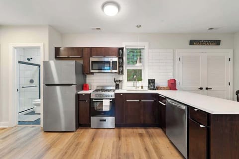 Modern kitchen with stainless steel appliances, dark wood cabinets, white countertops, and subway tile backsplash. Includes a coffee station, red toaster, and warm wood flooring