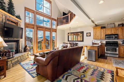 Cozy living room and kitchen with large windows, leather sofas, a stone fireplace, wooden cabinets, and vibrant rugs.