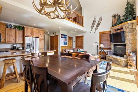 Rustic living room and kitchen area with wooden furniture, antler chandelier, mounted skis, and stone fireplace with TV.
