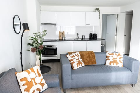 This chic living space features a pair of comfortable gray sofas, softened with warm-toned throws and patterned cushions for a welcoming touch. A minimalist white coffee table, elegant floor lamp, and carefully chosen décor elements—including lush potted plants and a modern wall mirror—add to the stylish appeal. Expansive windows bring in ample natural light, highlighting the a...