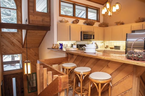 A cozy kitchen with wooden accents, three bar stools at a counter, white cabinets, and stainless steel appliances. Stairs lead down beside the kitchen. Large windows provide natural light.