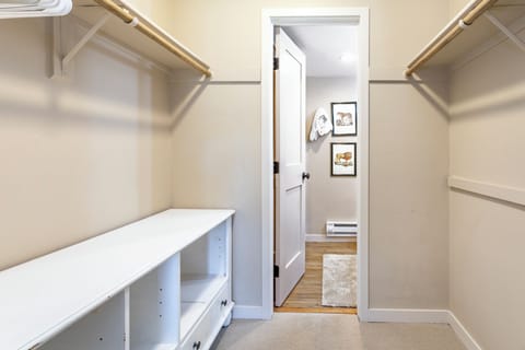 A walk-in closet with beige walls, empty wooden shelves and a white door leading to another room. The floor is carpeted.
