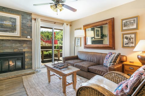 A cozy living room features a leather sofa, a wicker chair, a wooden coffee table, and a fireplace. A large mirror and framed photos adorn the walls. Sliding glass doors lead to an outdoor patio.