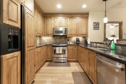 A modern kitchen with wooden cabinets, stainless steel appliances, granite countertops, and recessed lighting. The space includes a refrigerator, oven, microwave, dishwasher, and a double sink.