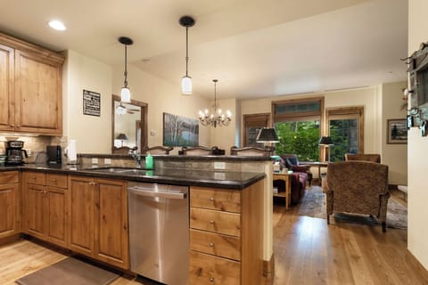A modern kitchen with wooden cabinetry, stainless steel appliances, and a granite countertop opens into a living area with a chandelier, sofa, and large windows.