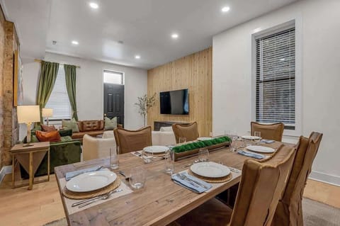 Lavish dining area with a wooden table and six cushioned chairs, featuring exposed brick and a colorful accent wall. Offers a view of the living room with a 65" TV and large window, creating a warm, open atmosphere.