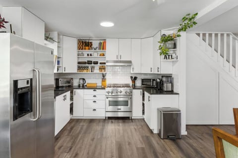 Modern kitchen with white cabinets, stainless steel appliances, tiled backsplash, and wooden floors. Plants and decor adorn the shelves and counter. Stairs are visible to the right.