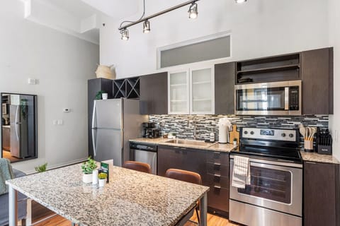 This modern kitchen features a spacious island with cabinetry, sleek white countertops, stainless steel appliances, open shelving, and stylish lighting.