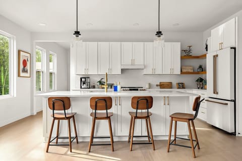 Sleek modern kitchen with white cabinetry, gold hardware, a spacious island with a farmhouse sink, pendant lighting, four wooden barstools, a stainless steel gas stove, a built-in refrigerator, open wooden shelves with decor, and a coffee station.