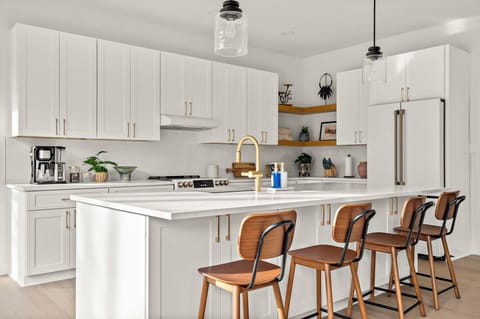 Sleek modern kitchen with white cabinetry, gold hardware, a spacious island with a farmhouse sink, pendant lighting, four wooden barstools, a stainless steel gas stove, a built-in refrigerator, open wooden shelves with decor, and a coffee station.