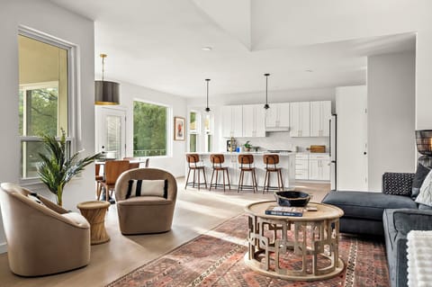 Stylish open-concept living space featuring a modern kitchen with white cabinetry, gold hardware, a large island with barstools, pendant lighting, a cozy living area with a plush sofa, accent chairs, a patterned rug, a round wooden coffee table, and large 