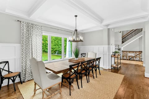 A bright and elegant dining room with a large farmhouse table, stylish seating, coffered ceilings, and a window offering scenic views of the backyard.