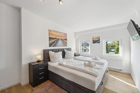 Das Schlafzimmer ist hell und freundlich eingerichtet, bietet ein großes Boxspringbett mit frischer Bettwäsche, zwei Nachttische mit Lampen, Wandbilder mit Strandmotiv sowie einen an der Wand montierten Fernseher.